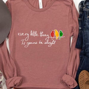 Puede incluir: Una camiseta de manga larga de color malva con el texto "every little thing is gonna be alright" en letras blancas. Tres pájaros coloridos están posados en un alambre por encima del texto.