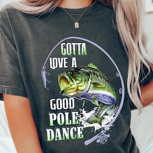 Könnte beinhalten: Ein dunkelgraues T-Shirt mit einem Grafikdruck eines Barsches, der aus dem Wasser springt, mit einer Angelrute und dem Text "Gotta love a good pole dance".