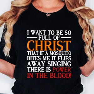 Può includere: Maglietta nera con testo bianco che recita "Voglio essere così pieno di Cristo che se una zanzara mi punge, vola via cantando che c'è potere nel sangue!"