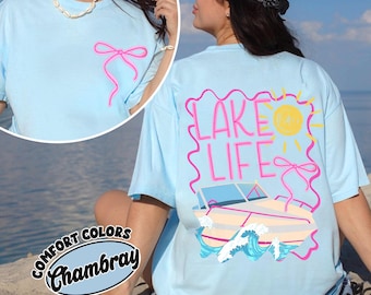 May include: A light blue t-shirt featuring a small pink bow graphic on the front left chest. The back displays a large graphic with the text "LAKE LIFE," a yellow sun, a pink bow, a boat, and blue waves, all enclosed in a pink wavy border.