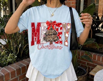 May include: A light blue short-sleeve t-shirt featuring the text MERRY in red and white patterns, with a leopard print Christmas tree graphic replacing the letter R, and the word Christmas in red script below the tree, paired with a white skirt.