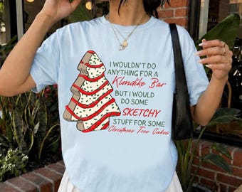 May include: A light blue t-shirt featuring a graphic of a Christmas tree cake with red icing and green sprinkles. The text reads: I WOULDN'T DO ANYTHING FOR A Klondike Bar BUT I WOULD DO SOME SKETCHY STUFF FOR SOME Christmas Tree Cakes.