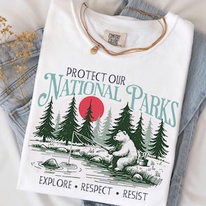 May include: White t-shirt with a graphic design promoting national parks. The design features a bear fishing in a river, pine trees, and a red sun. Text reads "PROTECT OUR NATIONAL PARKS" and "EXPLORE • RESPECT • RESIST".