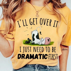 Puede incluir: Camiseta amarilla con una vaca de dibujos animados y el texto "I'll get over it I just need to be dramatic first."