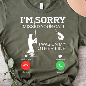 May include: Green t-shirt with a white graphic of a fisherman holding a fishing rod with a fish on the line. The text reads "I'm sorry I missed your call. I was on my other line." Below the graphic are two phone icons, one with a red decline button and the other with a green accept button.