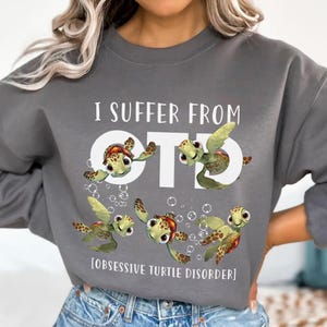 Puede incluir: Una sudadera gris con el texto "I suffer from OTD [OBSESSIVE TURTLE DISORDER]" y varias tortugas de dibujos animados impresas en ella.