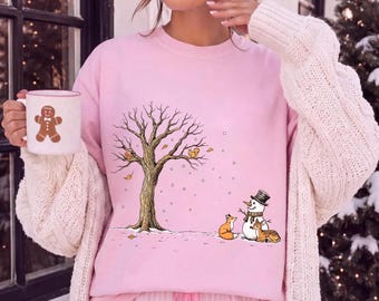 May include: A light pink crewneck sweatshirt featuring a graphic of a winter scene with a large tree, a snowman, a fox, and a deer. The sweatshirt is paired with a white mug that has a brown gingerbread man illustration printed on the front.