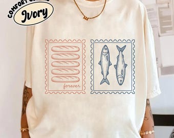 May include: An ivory t-shirt featuring two rectangular stamp-style graphics. The left stamp contains five stacked baguette illustrations with the word "forever" below them. The right stamp displays two fish illustrations. A "Comfort Colors Ivory" logo is visible.