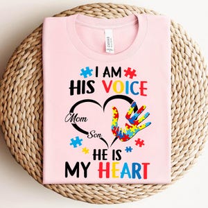 May include: Pink t-shirt with a heart design featuring a handprint with puzzle pieces. The text reads "I am his voice, Mom Son, He is my heart".