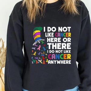 Puede incluir: Sudadera negra con un gráfico colorido del Gato en el Sombrero con una cinta. El texto dice "No me gusta el cáncer aquí o allá No me gusta el cáncer en ningún lugar".