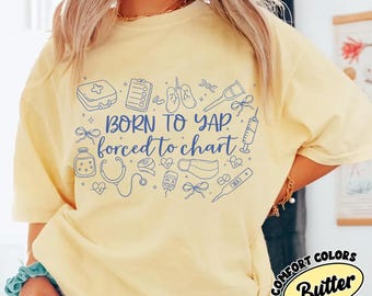 May include: A pale yellow t-shirt featuring a dark blue graphic print with the text "BORN TO YAP forced to chart" surrounded by various medical icons including a stethoscope, syringe, lungs, clipboard, thermometer, and bandages.