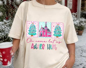 May include: An ivory short-sleeve t-shirt featuring a graphic print of two teal Christmas trees flanking a pink church building. Below the illustration, the text reads "Oh come let us Adore Him" in pink and teal cursive and block lettering.