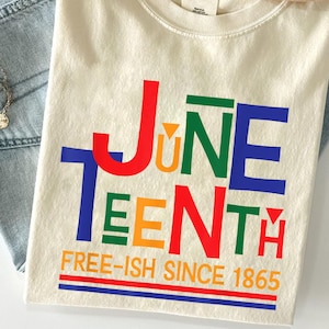 Pode incluir: Camiseta creme com a palavra "Juneteenth" em letras coloridas. O texto "FREE-ISH SINCE 1865" está abaixo, com listras vermelhas, laranja e azuis. A camiseta está dobrada sobre jeans azul.