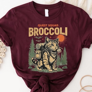 Könnte beinhalten: Burgunderfarbenes T-Shirt mit einer Grafik einer Katze mit Rucksack und Halstuch sowie dem Text "QUEST SQUAD BROCCOLI". Das Design umfasst eine Sonne, Bäume und Fledermäuse. Das Shirt ist aus weichem Material.