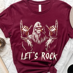 Op de afbeelding: Bordeauxkleurig T-shirt met een afbeelding van Bigfoot met zijn handen in de lucht, de rock-and-roll handgebaar makend. De tekst "Let's Rock" staat onder de afbeelding.