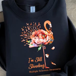 May include: Black sweatshirt with a pink flamingo standing on a flower. The flamingo is surrounded by orange ribbons. The text "I'm Still Standing" and "Multiple Sclerosis Awareness" is printed on the sweatshirt.