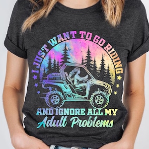 May include: A dark grey t-shirt with a colourful graphic design. The graphic features a woman driving a side-by-side vehicle in a forest setting. The text reads "I just want to go riding and ignore all my adult problems".