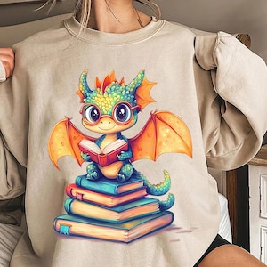 May include: A light beige sweatshirt with a cartoon illustration of a green dragon wearing glasses and reading a red book. The dragon is sitting on a stack of colorful books.