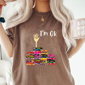 May include: Brown t-shirt with the text "I'm Ok" above a stack of colorful books. The graphic includes a yellow hand with the fingers pointing up. The books are in various colors, including red, blue, and green.
