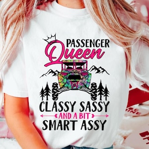 May include: White t-shirt with a pink and black graphic design. The design features a crown, the words "Passenger Queen", a colorful off-road vehicle, trees, and the text "Classy Sassy and a bit Smart Assy".