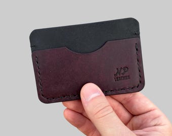 May include: A bi-colour leather cardholder. The top section is black, and the bottom is a deep burgundy. The cardholder has a stitched border and the initials "NP Leather" are embossed on the front. The cardholder is designed to hold credit cards.