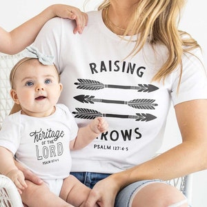 以下が含まれることがあります： 黒色の矢印のデザインと「PSALM 127:4-5」と「RAISING ROWS」の文字が書かれた、お揃いの白いTシャツを着た家族。Tシャツには「heritage of the LORD」の言葉が書かれている。家族が写真撮影のためにポーズをとっている。The Painted Porchはブランド名です。