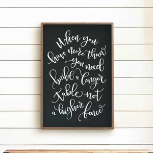 May include: A framed black chalkboard with white cursive text that reads "When you have more than you need, build a longer table not a higher fence." The frame is brown, and the sign hangs above a wooden cabinet.
