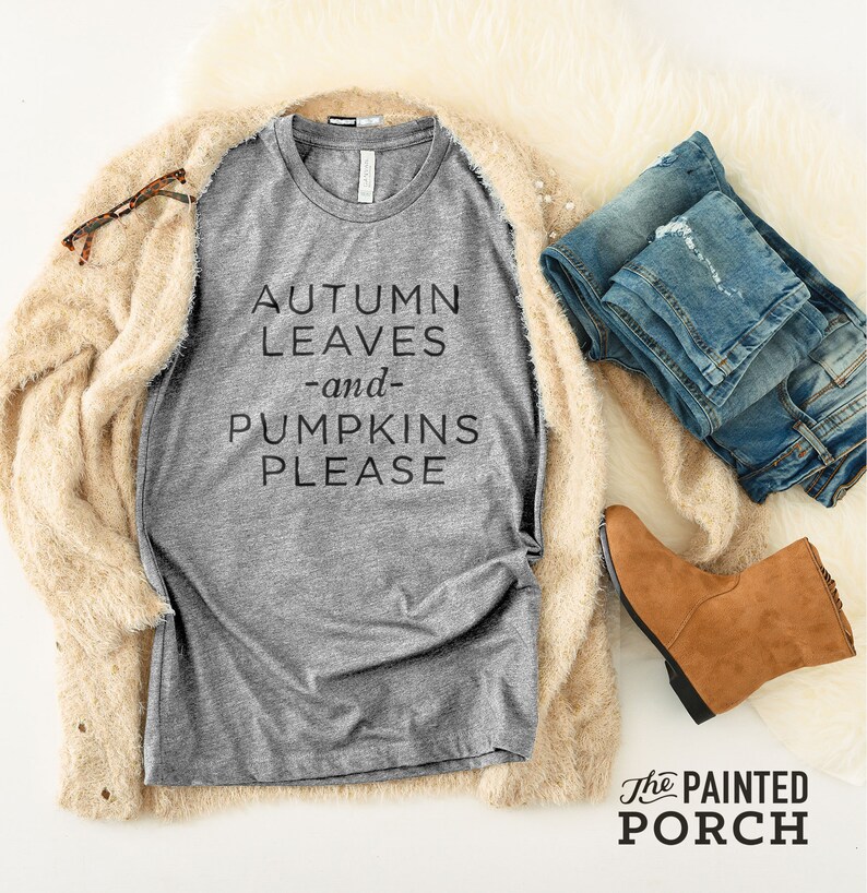 Pode incluir: Uma camiseta cinza com o texto "AUTUMN LEAVES -and- PUMPKINS PLEASE" impresso nela. A camiseta est&aacute; colocada sobre uma superf&iacute;cie branca com um casaco marrom, um par de cal&ccedil;as jeans azuis e um par de botas marrons.
