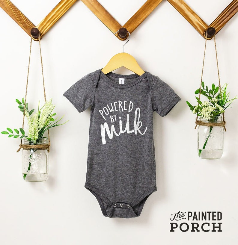 May include: A grey baby onesie with white text that reads "Powered by milk". The onesie is hanging on a wooden hanger against a white wall.