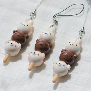 May include: Three decorative phone charms featuring a skewer of three cute animal-shaped treats. The top charm is a white bunny, the middle is a brown bear, and the bottom is a white bear on a stick. Each charm has a silver-colored metal loop and a string.