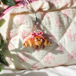 Könnte beinhalten: Ein bezaubernder Croissant-Anhänger mit einem lächelnden Gesicht, Rouge und einer rosa Schleife. Das Miniaturgebäck hat braune Details und ist an einem silberfarbenen Schlüsselanhänger befestigt. Der Hintergrund ist ein gesteppter Stoff mit rosa Schleifenmuster.