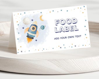May include: A folded white card with a watercolor illustration of a rocket ship, moon, and planets. The card reads "FOOD LABEL" and "ADD YOUR OWN TEXT" in blue lettering. The card is decorated with blue and gold stars.