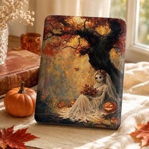 May include: A rectangular e-reader cover featuring a printed illustration of a skeletal figure in a white wedding dress and veil sitting under a dark tree. The scene includes orange pumpkins, autumn leaves, and warm golden light in the background.