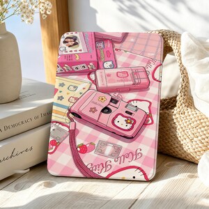Könnte beinhalten: Eine rechteckige Tablethülle mit einem rosa-weiß karierten Muster und Hello Kitty-Grafiken, darunter eine rosa Kamera, eine Schachtel und Erdbeer-Illustrationen. Das Design enthält den Schriftzug Hello Kitty in Schreibschrift.