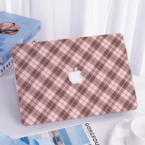 May include: A laptop case with a pink and brown plaid design. The case has a white Apple logo in the centre. The case is on a white surface, with a book and a magazine visible in the background.