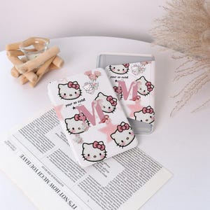 Könnte beinhalten: Weiße Handyhüllen mit einem sich wiederholenden Muster einer Zeichentrickkatze mit rosa Schleife, rosa Sternen und dem Buchstaben "M". Die Hüllen haben auch den Text "your so cute!".