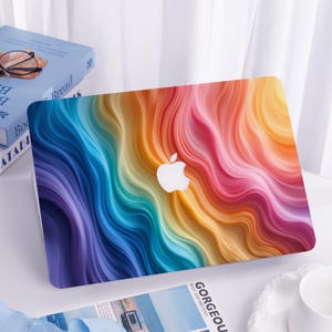 Może przedstawiać: Laptop z żywym, kolorowym wzorem. Pokrywa ma płynny, abstrakcyjny wzór w odcieniach niebieskiego, fioletowego, czerwonego, pomarańczowego i żółtego. Białe logo Apple jest wyśrodkowane. Laptop leży na białej powierzchni.
