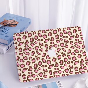 Könnte beinhalten: Eine Laptop-Hülle mit cremefarbenem Hintergrund und einem sich wiederholenden Muster aus braunen und rosa Leopardenflecken. Die Hülle hat einen Ausschnitt für das Apple-Logo. Bücher und Brillen sind im Hintergrund zu sehen.
