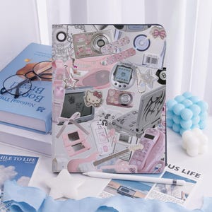 Puede incluir: Una funda blanca y rosa para tableta con un collage de electrónica retro y diseños de Hello Kitty. La funda incluye imágenes de cámaras, un Tamagotchi, un teléfono plegable y una consola de juegos. También se ven un lápiz óptico blanco y un libro azul.