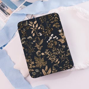 Könnte beinhalten: Ein rechteckiges, dunkelblaues Objekt mit einem botanischen Muster aus goldenen und weißen Blumen und Blättern. Das Design zeigt verschiedene Blattformen und kleine weiße Blüten, wodurch eine natürliche, elegante Ästhetik entsteht.