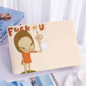 Könnte beinhalten: Eine hellbeige Laptop-Hülle mit einer Cartoon-Illustration eines Mädchens in einem orangefarbenen Kleid, das den Mittelfinger zeigt. Der Text "Fuck U" steht über dem Mädchen. Ein weißes Apple-Logo ist sichtbar.