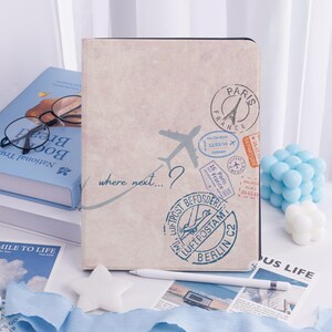 May include: A beige journal with travel-themed stamps and an airplane illustration. The cover features the text "where next...?" and various destination stamps. A white stylus and a star-shaped decoration are in the foreground.
