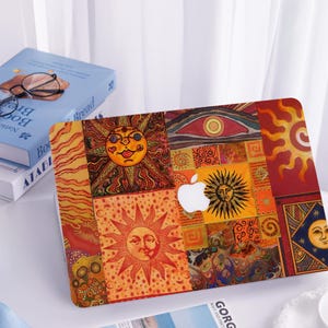 May include: A decorative laptop skin featuring a vibrant patchwork design with sun and moon motifs in shades of orange, red, and yellow. The Apple logo is visible. The design has a bohemian, artistic style.