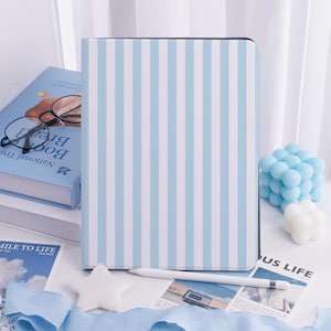 Op de afbeelding: Een tablet hoesje met een lichtblauw en wit gestreept patroon. Een witte styluspen ligt op een wit oppervlak. Een blauw boek met een bril en een blauwe decoratieve kubus zijn ook zichtbaar.