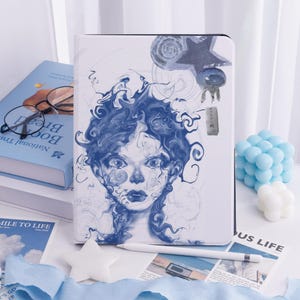May include: White tablet case featuring a blue and white illustration of a woman's face. A white stylus and a blue book are also visible. The case has a magnetic closure. The overall theme is art and design.