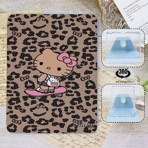 Könnte beinhalten: Eine Tablet-Hülle mit braunem Leopardenmuster und schwarzen Flecken und Schleifen. Die Hülle zeigt ein Cartoon-Bild von Hello Kitty auf einem Skateboard. Der Name "Mary" ist unten rechts in Kursivschrift geschrieben.