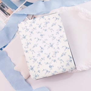 May include: A rectangular, floral-patterned notebook with a white background and blue flowers. The notebook has a dark blue border and is surrounded by light blue ribbons and a white star-shaped object.