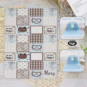 Op de afbeelding: Een tablet hoesje met een patchwork design, met strikken, taarten en "cute kitten" illustraties in lichtblauw, bruin en wit. De naam "Mary" staat op de hoes gedrukt. De hoes heeft ook een 360 graden rotatie functie.