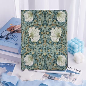 Könnte beinhalten: Eine Tablet-Hülle mit einem floralen Muster in Grün-, Creme- und Brauntönen. Das Design zeigt große weiße Blumen und komplizierte Blatt- und Stieldetails. Ein weißer Stift und ein Buch sind ebenfalls zu sehen.