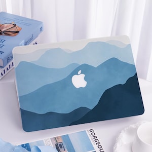 Könnte beinhalten: Eine blaue Laptop-Hülle mit einem Bergketten-Design in Blau- und Weißtönen. Das Apple-Logo ist in der Mitte sichtbar. Die Hülle liegt auf einer weißen Oberfläche mit einem Buch und einer Zeitschrift im Hintergrund.